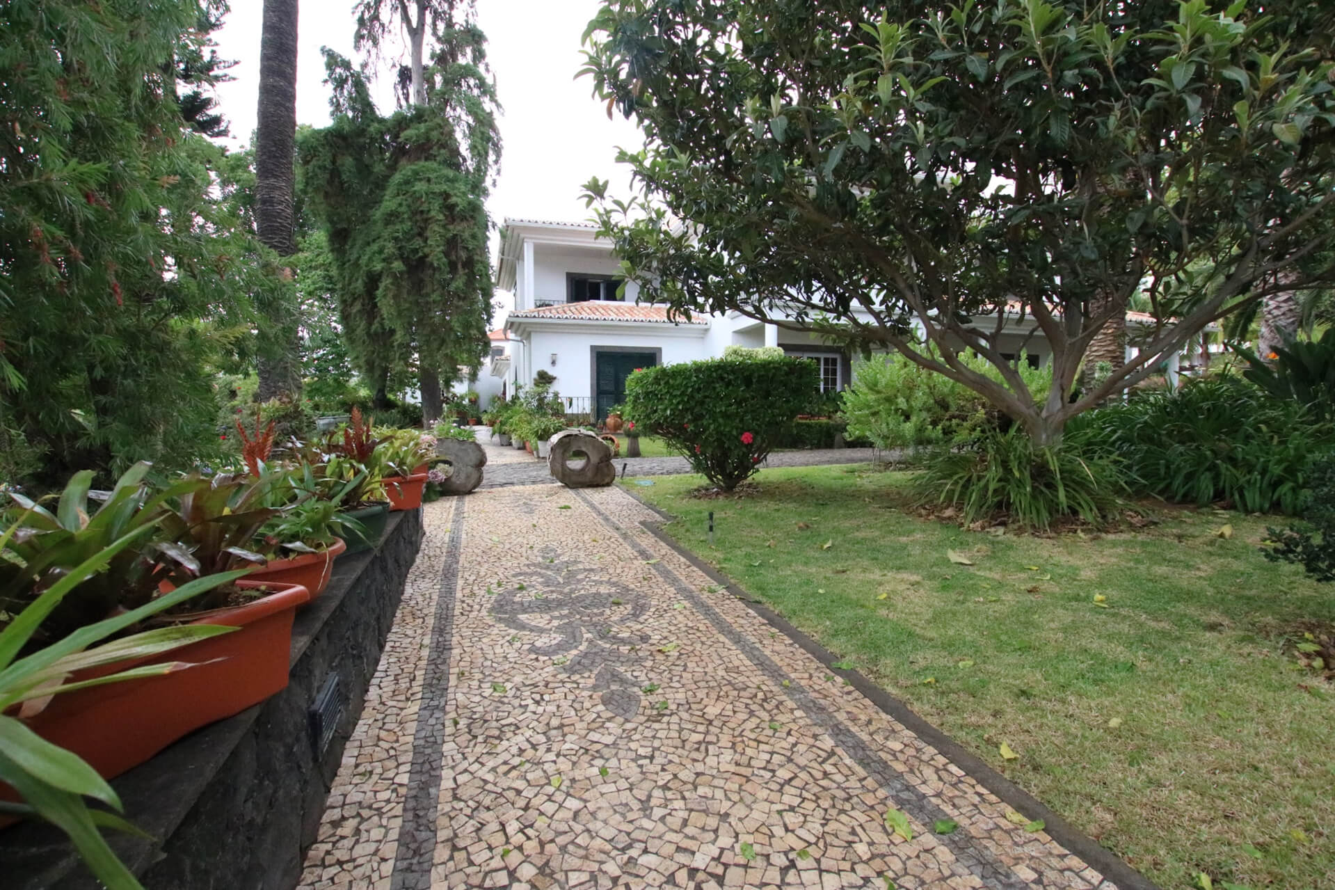 A walkway of the house with mosaic designs on it's floor with the garden next to it and the side facade of the property at the end of that walkway