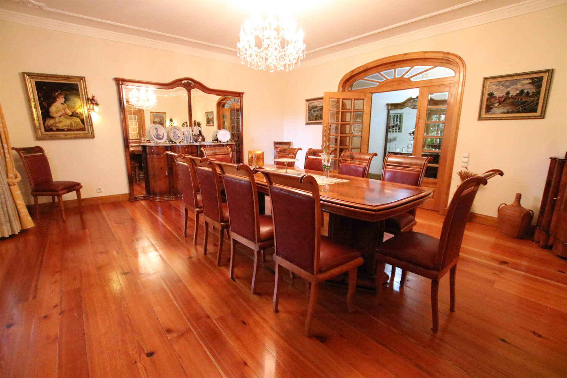 Dining room with a woodern floor a large table with chairs around it, some paintings on the wall and a mirror on top of a cupboard